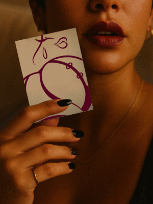 Close-up of a person holding the But A6 fine-art print by SKI; brown line art on textured paper, deep red lips, gold necklace and ring under warm lighting.