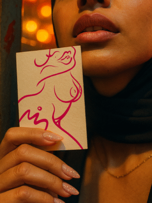 Close-up of the Veiled Queen A6 fine-art print by SKI held in hand; magenta line artwork on textured paper with bokeh city lights and veil/scarf details.